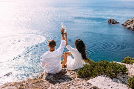 A couple in love on the shore drinking champagne, rear view. Vacation abroad. Holidays on the islands. A couple in light clothes is relaxing on the coast. Honeymoon. Wedding travel. Copy spaceの写真素材