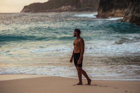 A man with a naked torso on the seashore. A lonely man stands on the beach. A man meets the sunset on the beach in Indonesia. Copy spaceの写真素材