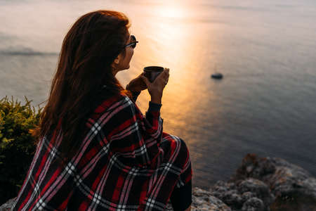 Young woman drinking tea from her flask, enjoying autumn evening by the sea. A woman meets the sunset on the sea. Autumn outdoor recreation. A young woman drinks hot tea on the coastの写真素材