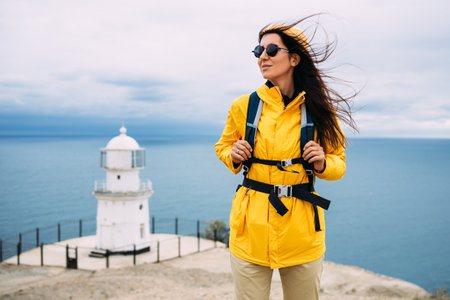 A traveler on the background of a lighthouse by the sea. Portrait of a traveler in yellow tourist clothes on the coast. A traveler by the ocean. Portrait of a girl traveling with a backpackの写真素材