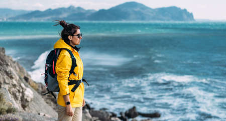Tourist with a backpack against the sea, panorama. Portrait of a woman in tourist gear against the sea. Travel and active lifestyle concept. Portrait of a traveler in a yellow jacket. Copy spaceの写真素材