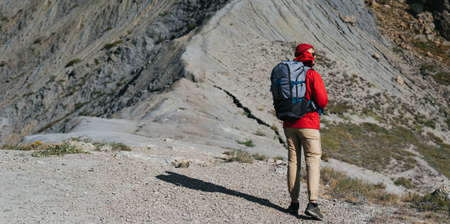 A male tourist with a backpack climbs the mountains, panorama. A traveler on the background of mountains, rear view. A tourist with a backpack on the background of mountains. Copy spaceの写真素材