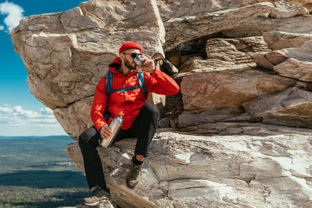 A male tourist is resting after a hard climb up the mountain. A tourist drinks tea from a flask in the mountains. A male traveler drinks hot coffee from a flask in the mountains. Copy spaceの写真素材