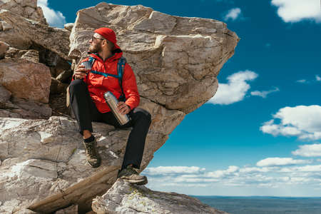A male tourist is resting after a hard climb up the mountain. A tourist drinks tea from a flask in the mountains. A male traveler drinks hot coffee from a flask in the mountains. Copy spaceの写真素材