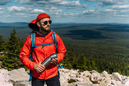 A bearded traveler with a backpack on the top of a mountain. Portrait of a traveler in a red cap and sunglasses. A tourist with a backpack stands against the background of a mountains. Copy spaceの写真素材