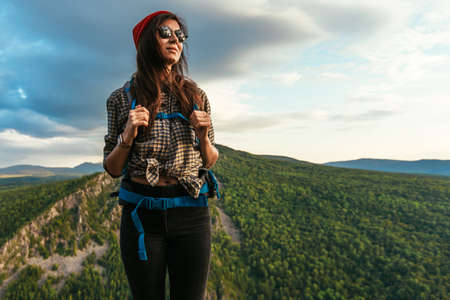A young tourist woman with a backpack enjoys the sunset from the top of the mountain. Portrait of a traveler in a red riding hood. A tourist with a backpack is standing on the top of the mountainの写真素材