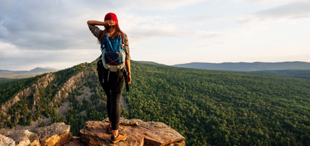 A young female tourist with a backpack admires the sunset from the top of the mountain. A traveler on the background of mountains. A traveler on the background of mountains at sunset, panoramic view.の写真素材