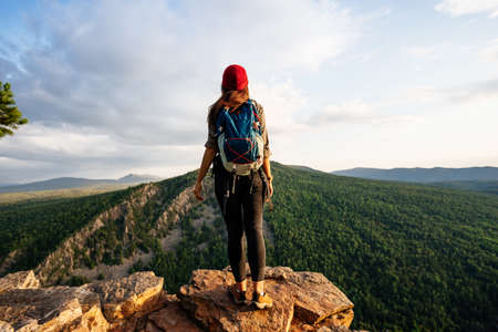 A young female tourist with a backpack admires the sunset from the top of the mountain. A traveler on the background of mountains. A traveler on the background of mountains at sunset. Copy spaceの写真素材