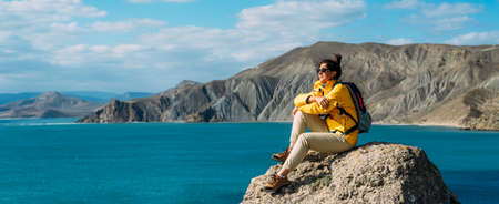 Tourist with a backpack against the sea, panorama. Portrait of a woman in tourist gear against the sea. Travel and active lifestyle concept. Portrait of a traveler in a yellow jacket. Copy spaceの写真素材