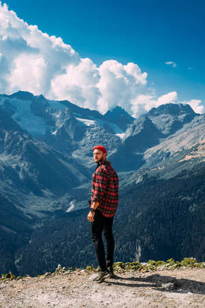 Stylish bearded man on top of a mountain. Portrait of a traveler in a red cap and a plaid shirt against the background of mountains. The concept of recreation and tourism. Travel. Copy spaceの写真素材