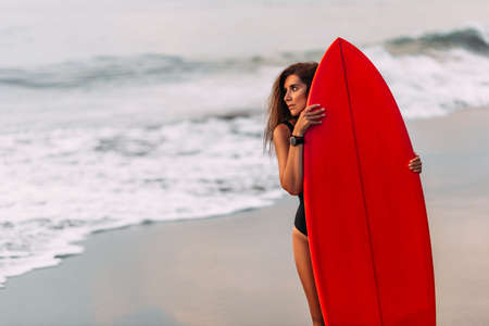 Beautiful sexy surfer girl on the beach at sunset. Beautiful girl with a surfboard. Surfer girl. Attractive sexy young woman in black swimsuit is standing on beach with red surfboard in handsの写真素材