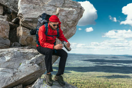 A bearded traveler with a backpack on top of a mountain. Portrait of a traveler in red clothes and sunglasses. A tourist with a backpack sits against the background of mountains. Copy spaceの写真素材
