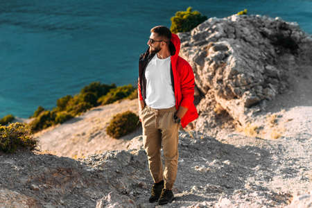 Portrait of a brutal bearded man in a red jacket by the sea. Male portrait on the background of a seascape. A traveler on the background of the sea coast. A man in a jacket at sunsetの写真素材