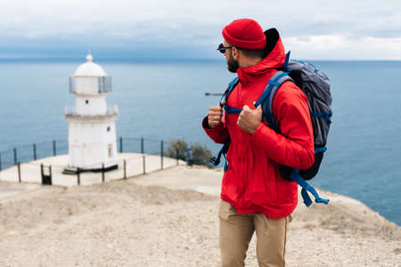 A traveler on the background of a lighthouse by the sea. Portrait of a traveler in red clothes on the coast. A traveler by the ocean. Portrait of a bearded man traveling with a backpack. Lighthouseの写真素材
