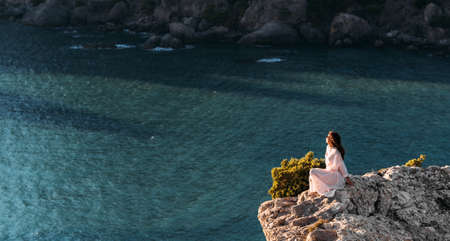 Beautiful girl sitting on a high rock and looking out to sea, panorama. Woman sits on the edge of cliff. The girl of the sea. Seascape. Woman at sunset. Sea tour. Copy spaceの写真素材