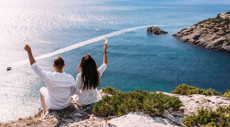 A man and a woman drink champagne on the seashore, rear view. Wedding trip to the sea, panorama. A couple in love in white clothes on the coast. Couple with champagne by the sea. Wedding travelの写真素材
