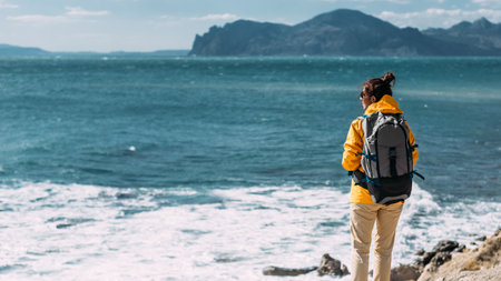 Tourist with a backpack against the sea, panorama. Portrait of a woman in tourist gear against the sea. Travel and active lifestyle concept. Portrait of a traveler in a yellow jacket. Copy spaceの写真素材