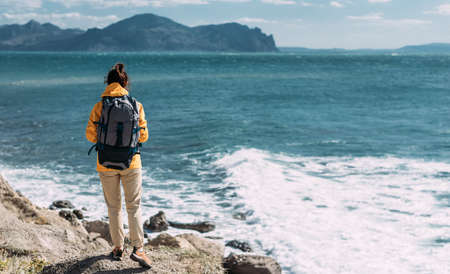 Tourist with a backpack against the sea, panorama. Portrait of a woman in tourist gear against the sea. Travel and active lifestyle concept. Portrait of a traveler in a yellow jacket. Copy spaceの写真素材