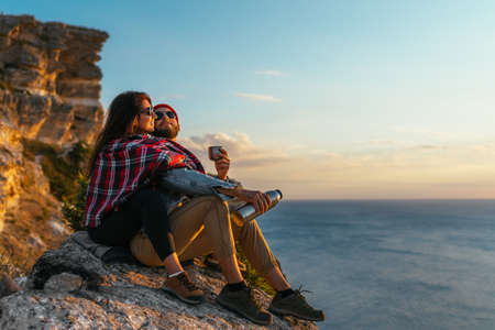 A stylish couple admires the seascape at sunset. A couple in love at sunset by the sea. A man and a woman are traveling. A beautiful couple meets the sunset on the seashore. Couple on the capeの写真素材