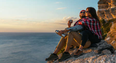 A stylish couple meets the sunset on the seashore. A beautiful couple at sunset by the sea. A couple in love admires a beautiful sunset on the coast. Lovers on the background of the sea. Copy spaceの写真素材
