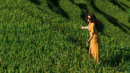 A beautiful woman walks on rice terraces on the island of Bali. A beautiful woman in a yellow dress walks through the green rice fields. A tourist traveling in Indonesia. copy spaceの写真素材
