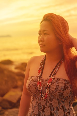 Portrait of an asian woman with hand holding the back of her head looking far away at beach on sunset.の写真素材
