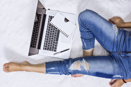 Woman wearing denim jeans sitting on bed and working with laptop. Domestic life lifestyle concept.の写真素材
