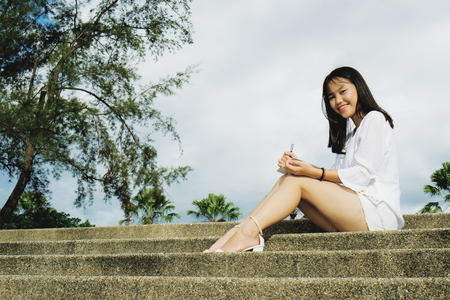 Smiling young Asian woman sitting on concrete stairs in a public park. Outdoor lifestyle photography.の写真素材