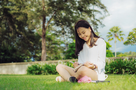 Smiling young Asian woman sitting with legs cross on grass field in a public park while writing on diary. Outdoor lifestyle photography.の写真素材