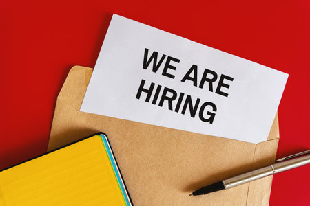 "Hiring Announcement on Red Background" - A white paper with a hiring announcement enclosed in an envelope, accompanied by office stationery, set against a bold red background, implying the excitement and significance of a new job opportunity.の写真素材