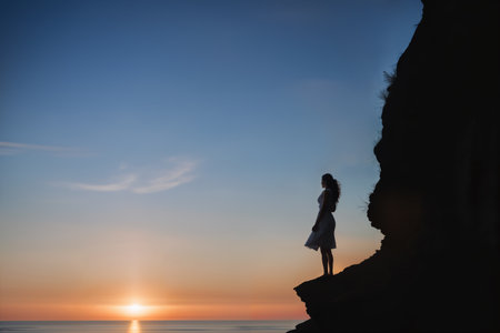 Solitude and tranquility. Woman standing on cliff, gazing at sunset or sunrise over the ocean. AI Generated Imageの素材