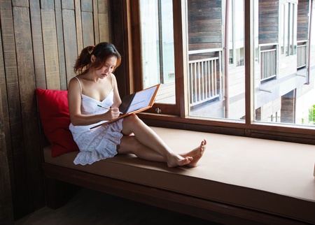 A woman in white dressing leaning on red pillow is reading a folder sitting near bay windowの写真素材