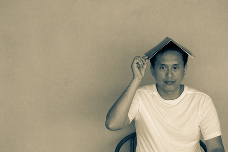 Sitting Asian man tipping his fingers on book on orange wallの写真素材