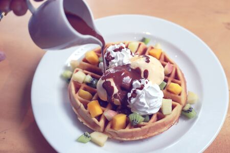 Chocolate waffle with ice cream and whipped cream and chocolate sacue toppig watering from top by small jar. Sweet cold European dessert on white plate.の写真素材