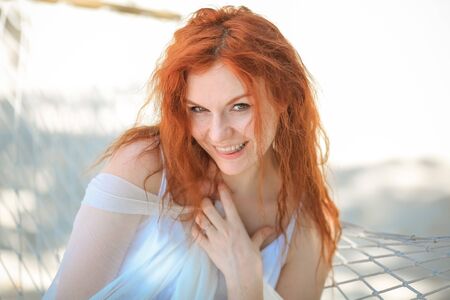 portrait of a red-haired girl with a happy smile in a white dress on the background of white sandの写真素材