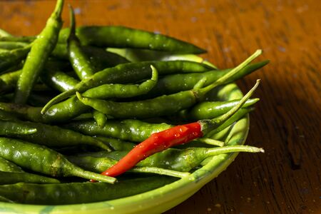 one red chilli among green chillies on a plate and wooden tableの写真素材