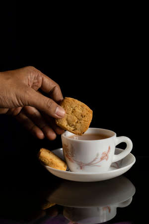 Portrait of human hand holding backed biscuit above tea cup plate on shinny reflecting surface with...