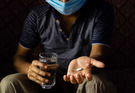 selective focus on a tablet or pill on palm of an indian person with nose mask on glass of water on other hand at homeの写真素材