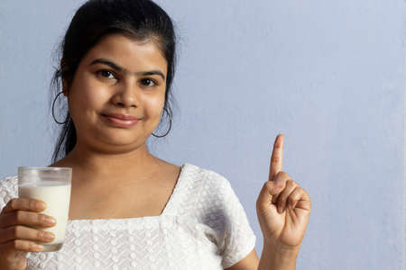 an Indian / asian woman holding a glass of milk with smile, looking at camera and pointing finger on...