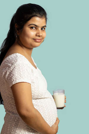 an Indian / asian pregnant woman holding glass of milk with smile on cyan backgroundの写真素材