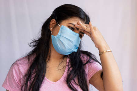 A mid adult Indian Asian woman wearing surgical nose mask and touching forehead for headache, health...