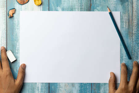 Top view of blank white paper page on blue wooden background with human hand, pencil, eraser, sharpenerの写真素材