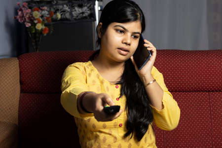 An Indian woman talking over phone while holding remote of television