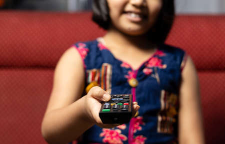An Indian girl child smiles while watching television with remote in hand and sitting on sofaの写真素材