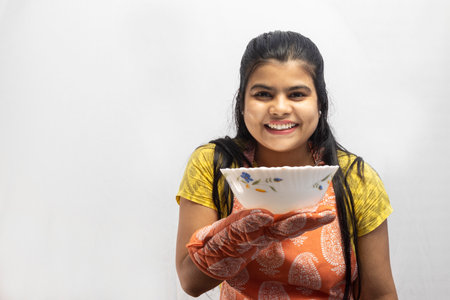 A pretty Indian housewife woman wearing cooking apron and oven gloves with a serving bowl in hand smiles on white backgroundの写真素材