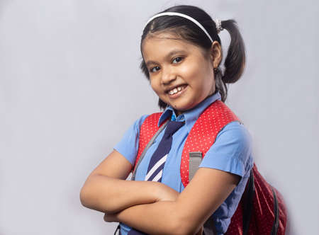 Portrait of a happy smiling Indian girl child student in blue school uniform with red bag on grey backgroundの写真素材