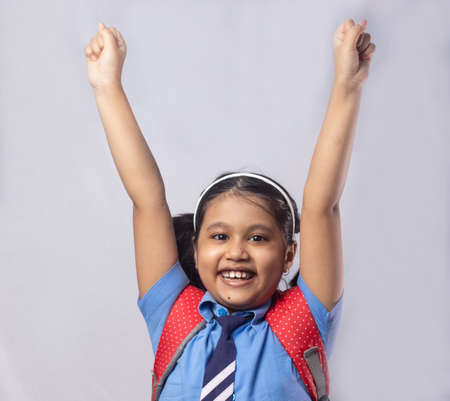 A happy cheerful smiling Indian girl child student in blue school uniform with red bag on grey backgroundの写真素材