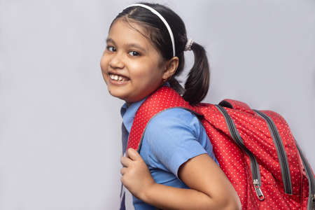 Side view of a happy smiling Indian girl child student in blue school uniform with red bag on grey backgroundの写真素材