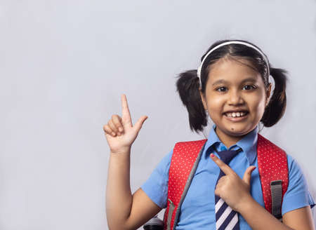 Portrait of a happy smiling Indian girl child student in blue school uniform with red bag pointing to blank area on grey backgroundの写真素材