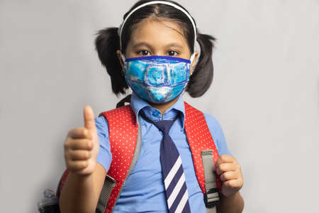 A happy Indian girl child student in blue school uniform with red bag and nose mask protection shows thumbs up on grey backgroundの写真素材